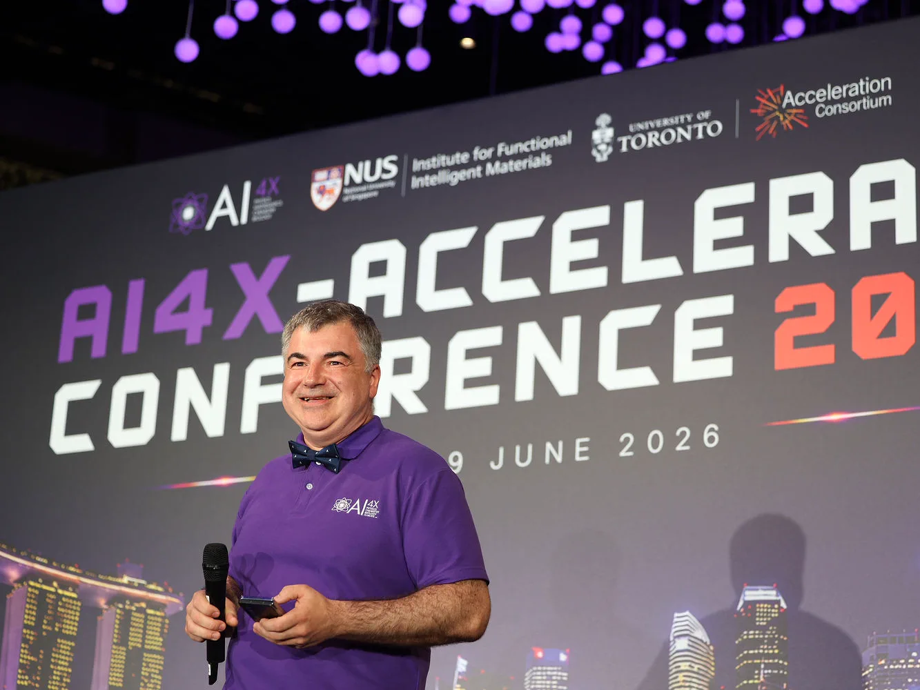 Speaker in a purple AI4X shirt on stage in front of the AI4X-Accelerate Conference 2026 backdrop